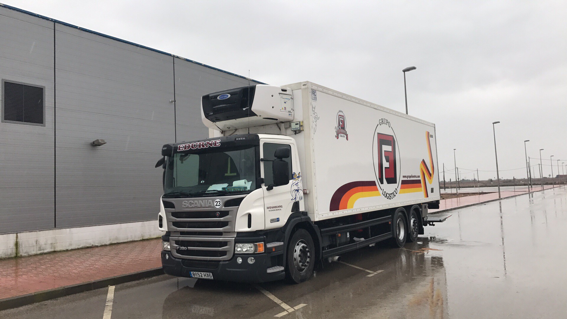 Camión refrigerado Scania P320 con la inscripción "EDURNE" en un día lluvioso en un estacionamiento industrial.